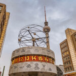Die Weltzeituhr am Alexanderplatz in Berlin mit dem Fernsehturm im Hintergrund bei bewölktem Himmel.