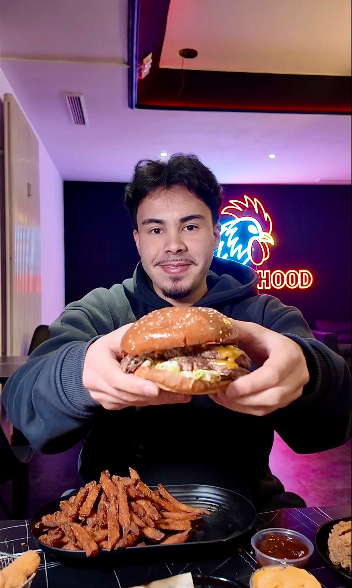 Mann hält lächelnd einen großen Burger im Chickenhood Datteln vor Neon-Logo