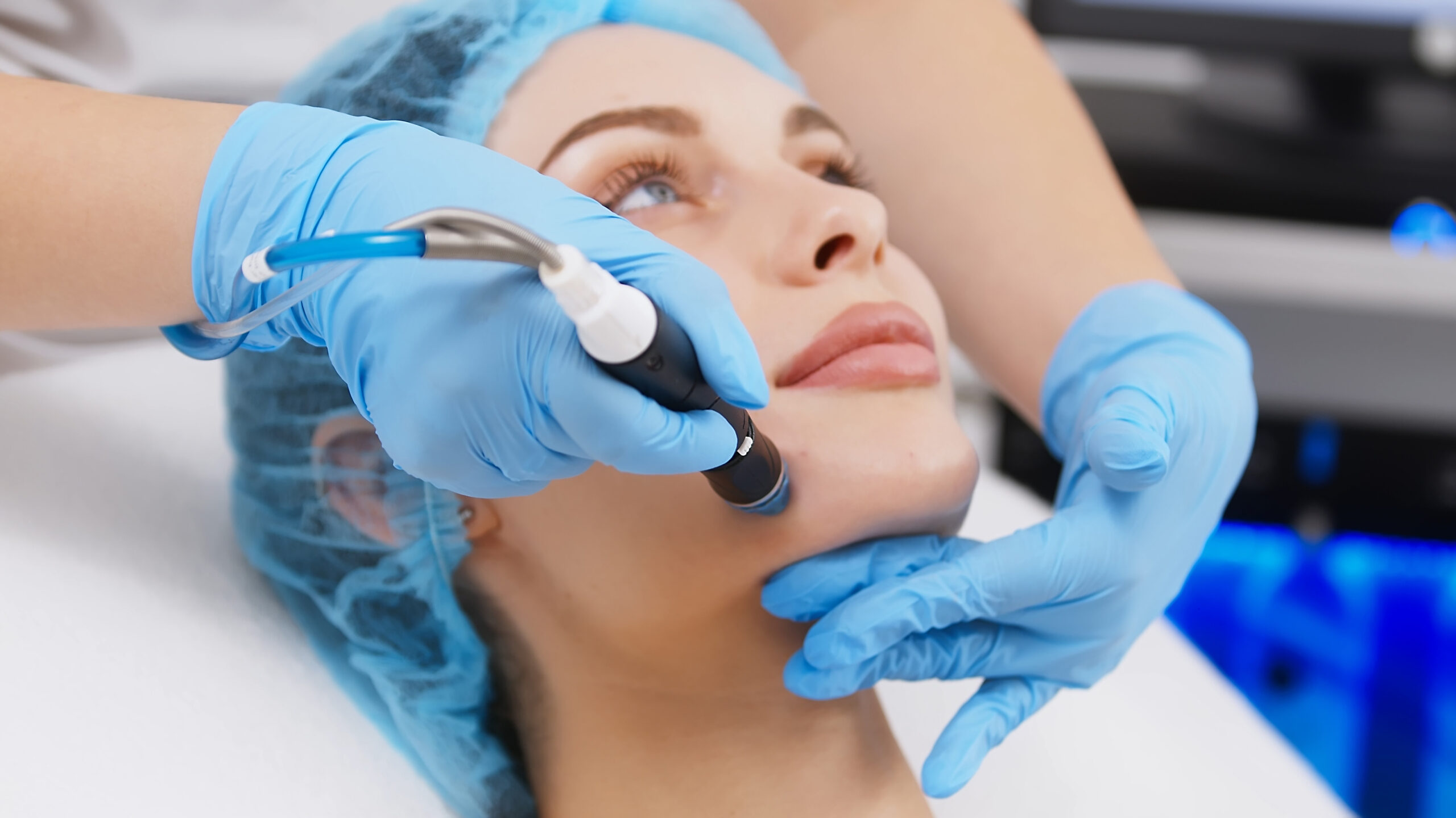 Nahaufnahme einer jungen Frau bei einer AquaFacial-Behandlung im Gesichtsbereich. Eine Kosmetikerin mit blauen medizinischen Handschuhen hält sanft das Gesicht der Frau und behandelt ihre Haut mit einem speziellen AquaFacial-Gerät.