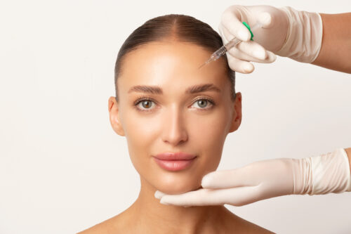 Frau mit glatter Haut blickt ruhig in die Kamera, während eine Fachkraft mit weißen Handschuhen eine Botulinum-Injektion in die Stirn vorbereitet