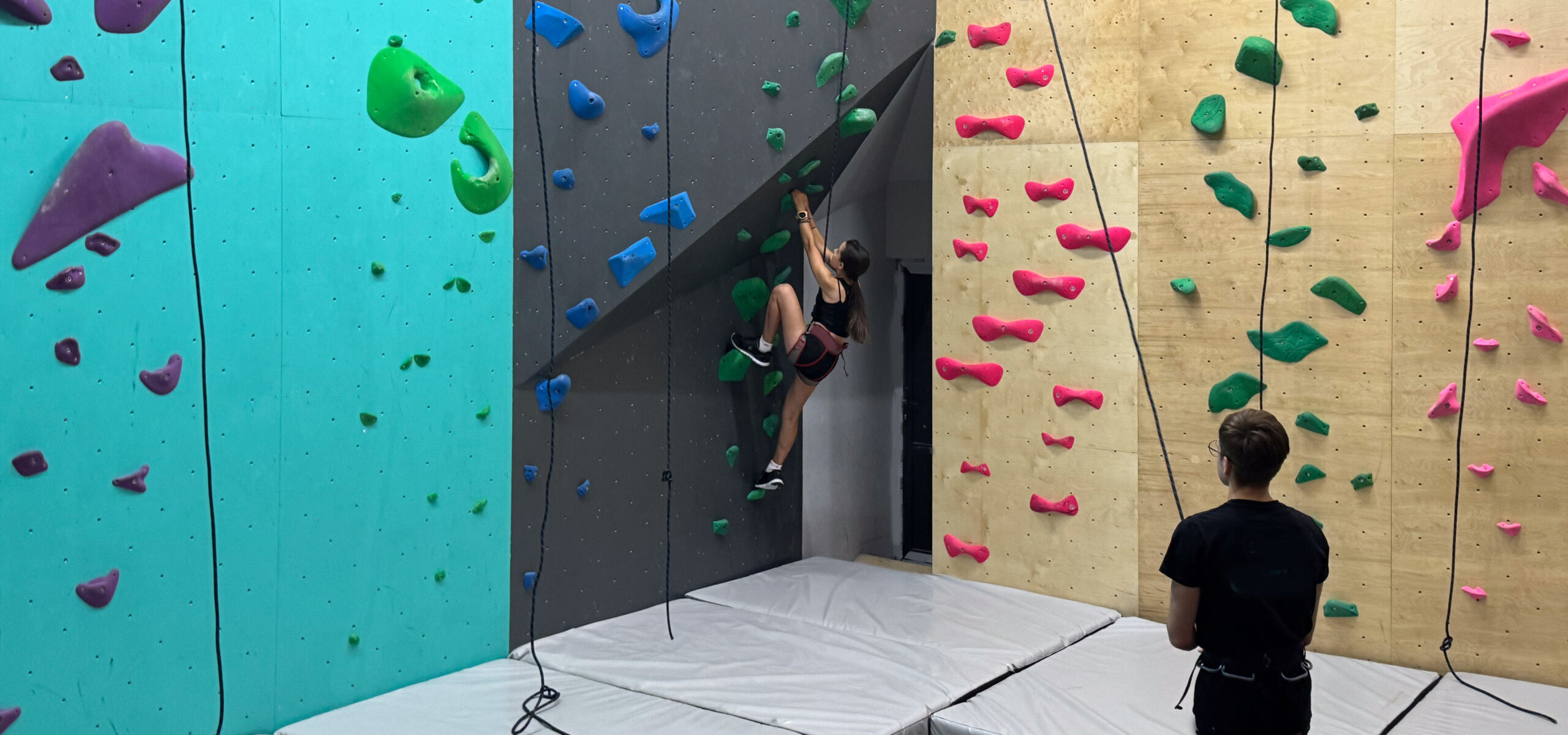Frau klettert an einer bunten Indoor-Kletterwand in der MOVE Kletterhalle mit Sicherungsseil und weichen Matten
