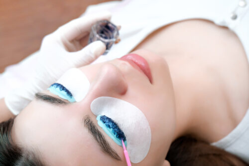 Nahaufnahme einer liegenden Frau während einer professionellen Wimpernfärbung. Ihre Augen sind geschlossen und mit Schutzpads versehen, während eine Kosmetikerin mit Handschuhen die Farbe sorgfältig auf die Wimpern aufträgt.