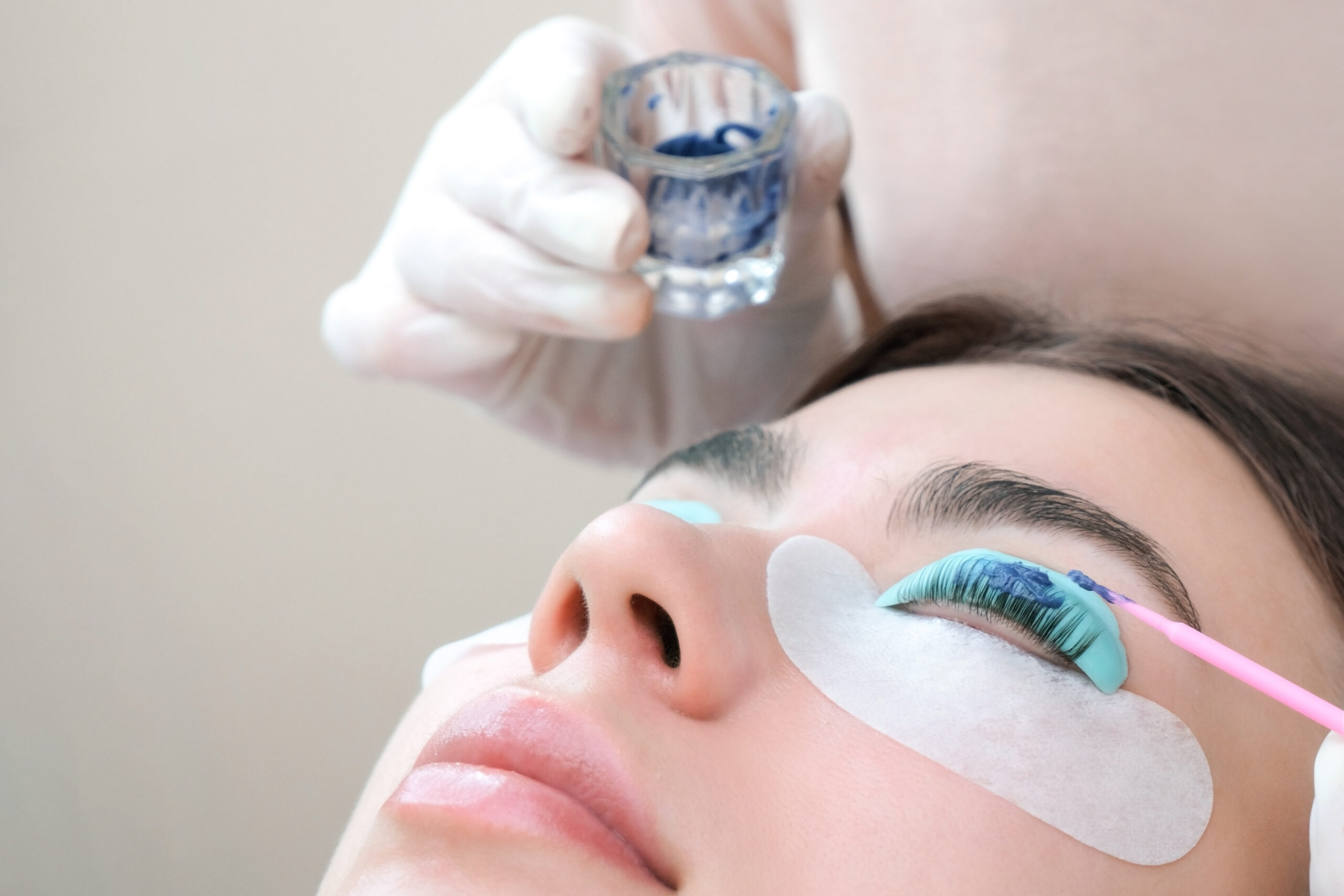 Nahaufnahme einer jungen Frau, die eine professionelle Wimpernfärbung erhält. Ihre unteren Augenlider sind mit Schutzpads bedeckt, während eine Kosmetikerin mit Handschuhen die Farbe sorgfältig aufträgt.