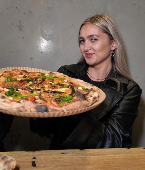 Frau präsentiert lächelnd eine frisch gebackene, großzügig mit Gemüse belegte Pizza bei Zampano To Go Frankfurt