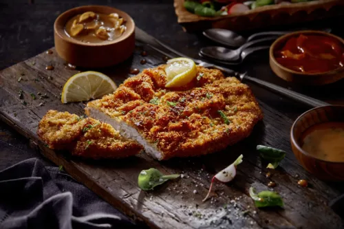 Knusprig gebratenes Schnitzel auf einem Holzbrett, garniert mit Zitronenscheiben, Radieschen und Kräutern, im Hintergrund Beilagen und Saucen