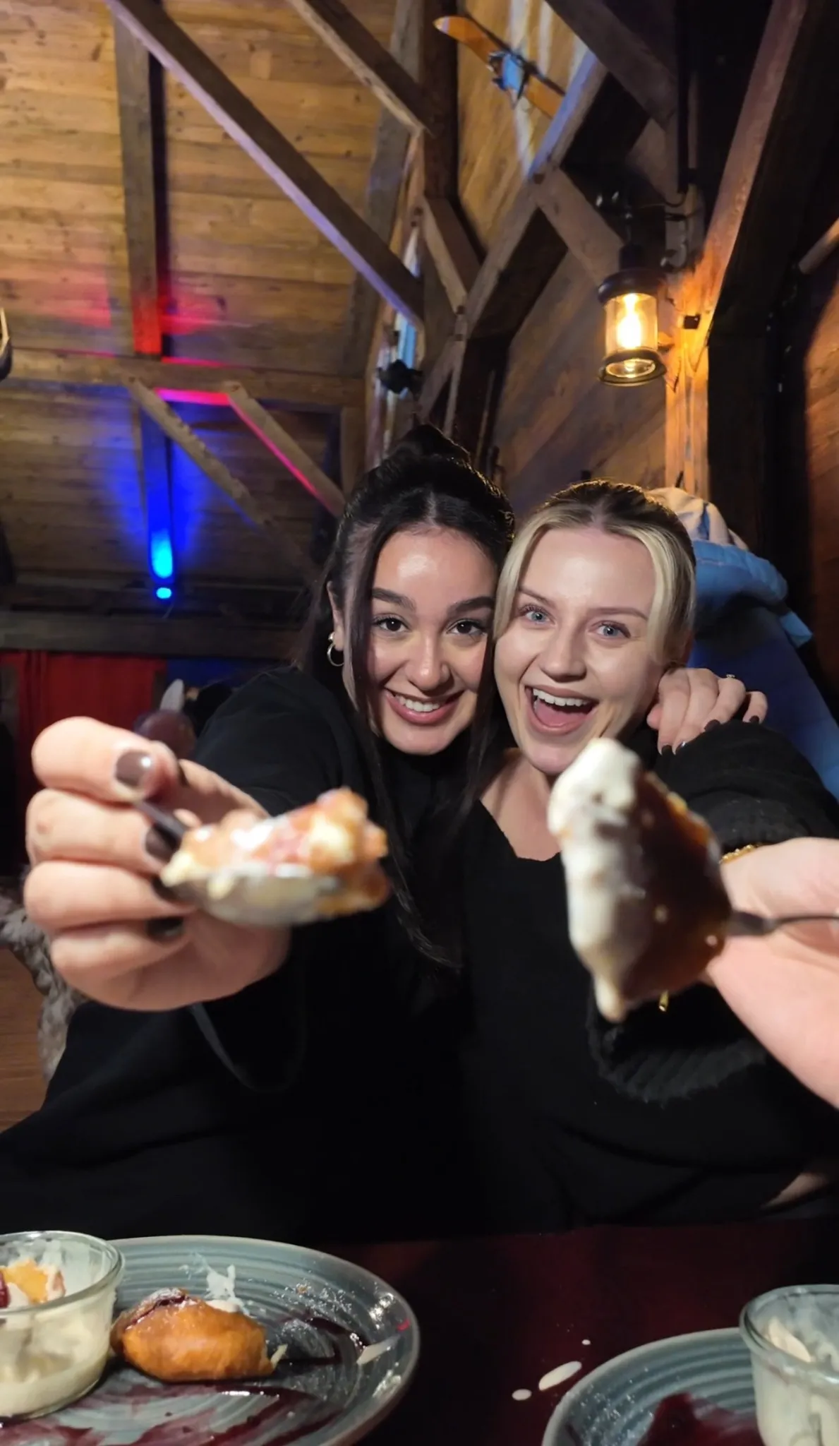 Zwei fröhliche Gäste in der Alm Fernweh halten Dessertstücke mit Sahne in die Kamera, im Hintergrund gemütlich-rustikale Almhüttenatmosphäre