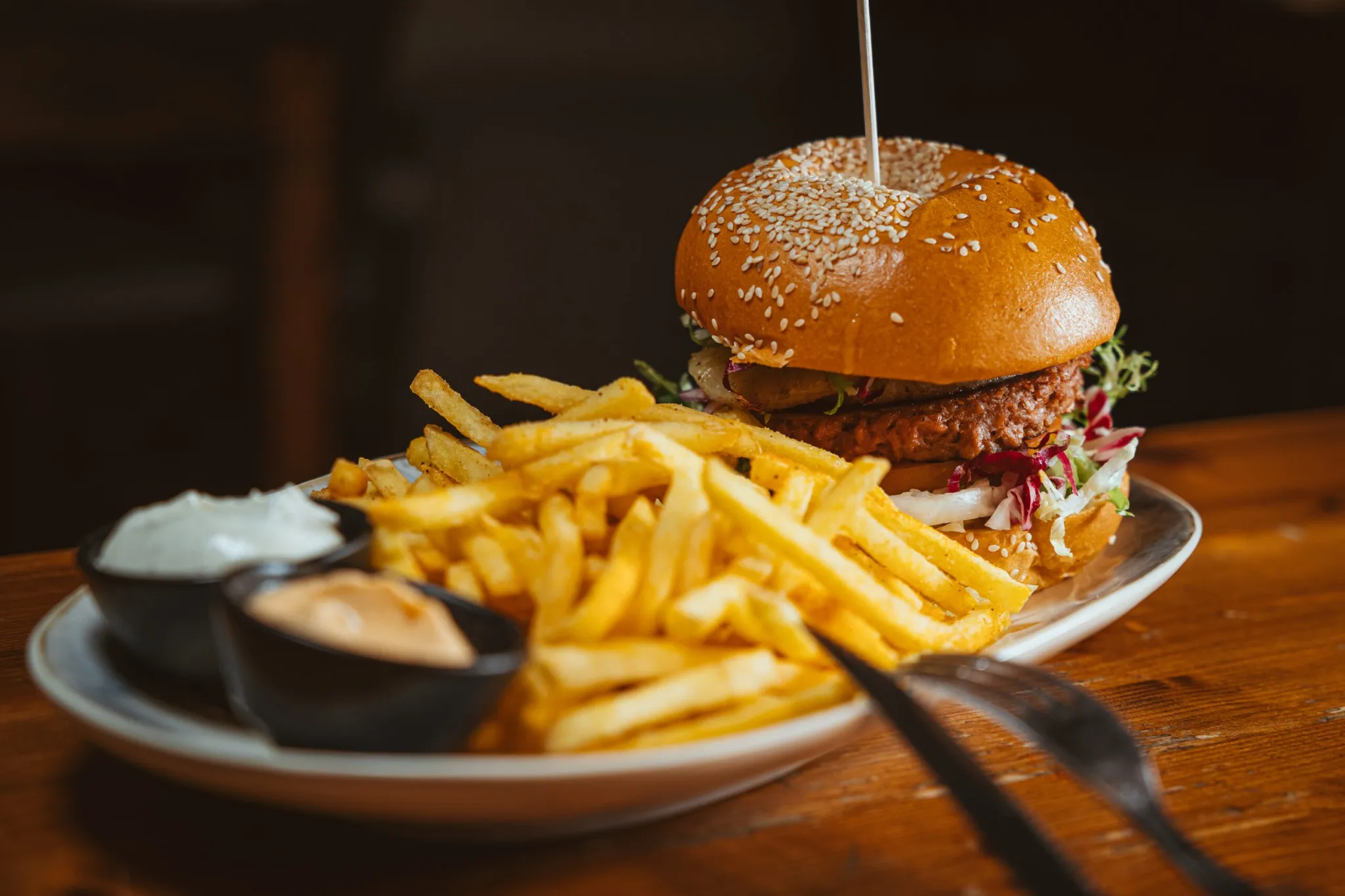 Saftiger Burger mit Sesam-Bun, serviert mit einer großen Portion Pommes frites und Dips auf einem ovalen Teller im Blondies Bochum