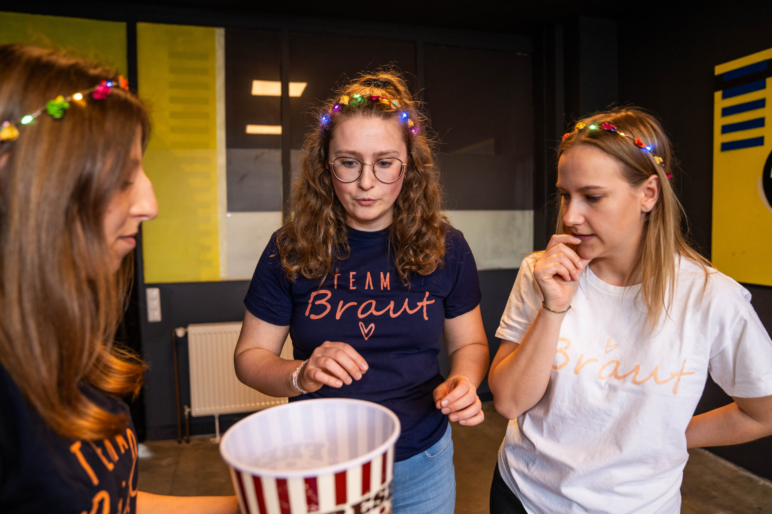 Drei junge Frauen beim Junggesellinnenabschied in der Face Off GmbH, eine trägt ein "Team Braut" Shirt, sie stehen vor einem Spiel mit Popcorn-Eimer