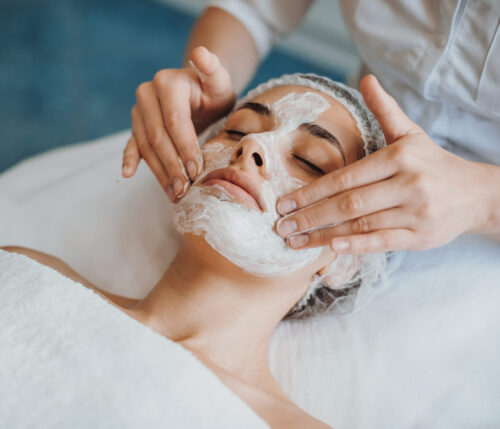 Frau liegt entspannt mit Maske im Gesicht, eine Kosmetikerin massiert die Maske sanft ein – Spa-Behandlung auf der Liege