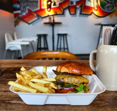 Burger mit Salat, Tomate, Gurke und Cheddar sowie Pommes in einer weißen To-go-Box auf Holztisch, im Hintergrund Graffiti-Wand im Hornochse Köln