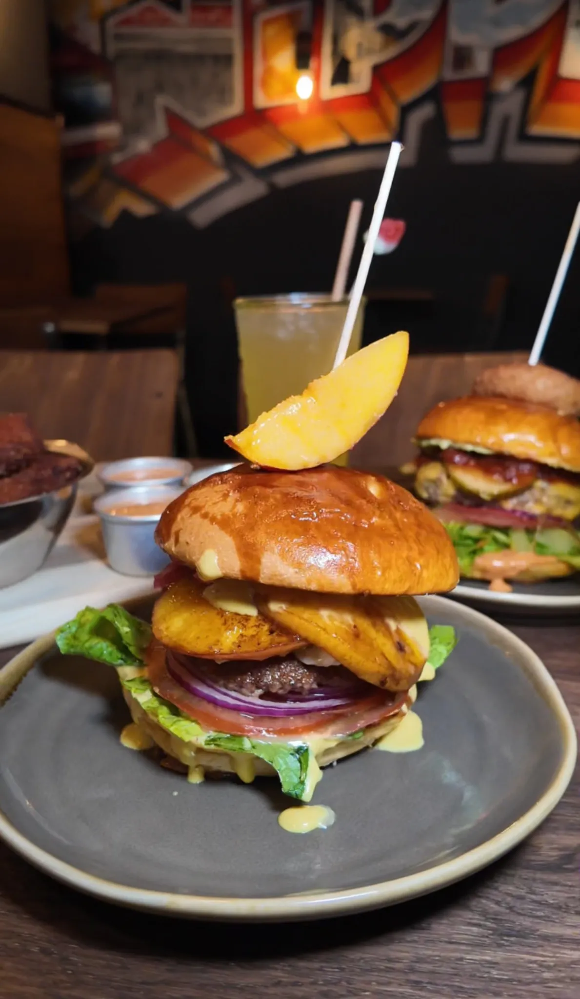 Burger mit gegrillter Ananas, Rindfleischpatty, roten Zwiebeln, Salat und Brioche-Bun, garniert mit Ananasschnitz, im Hintergrund Limonade und weitere Burger im Hornochse Köln