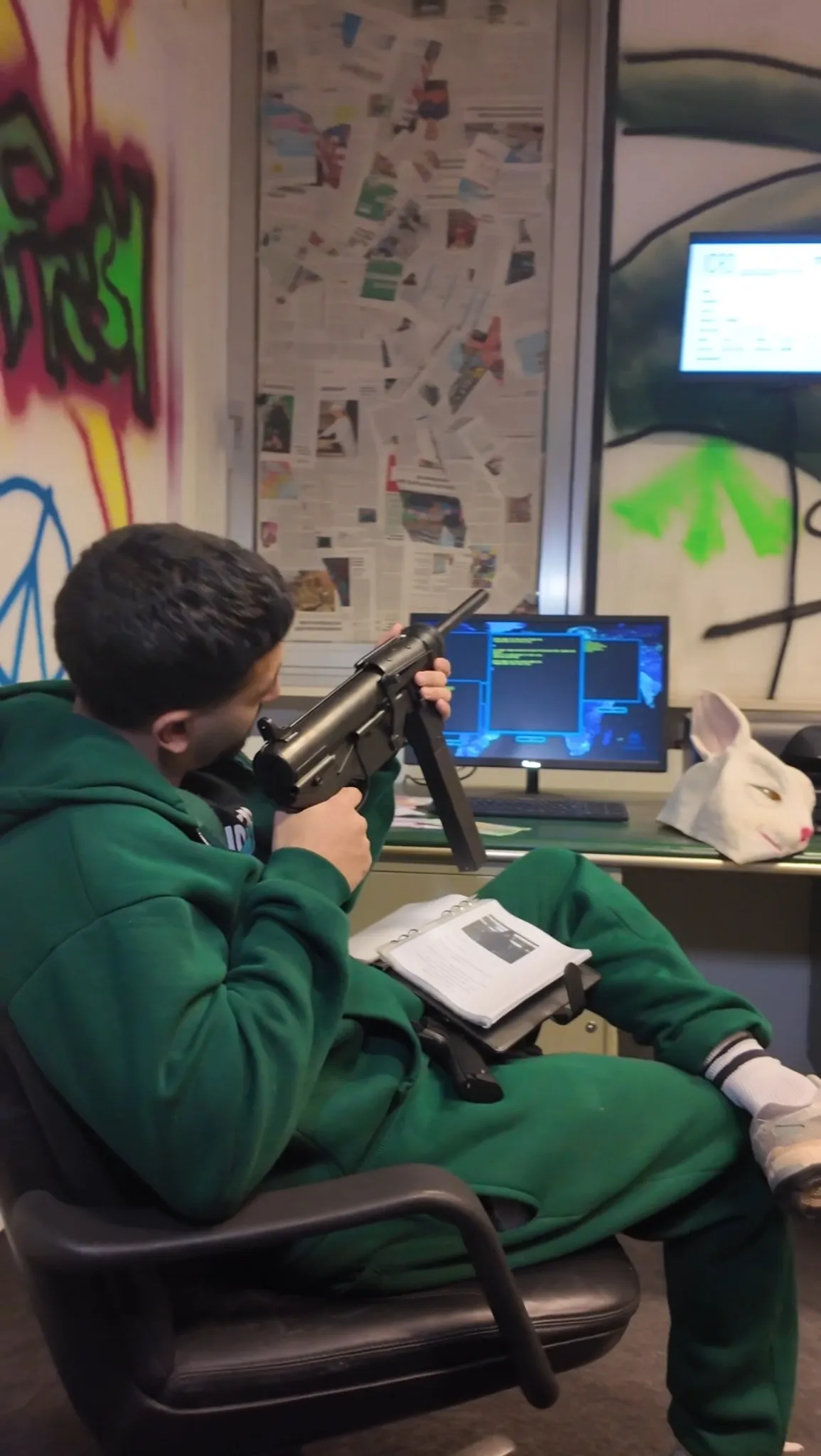 Teilnehmer in grüner Kleidung sitzt im Hacker-Raum bei Face Off Rüsselsheim mit Spielzeugwaffe, Buch, Hasenmaske und Bildschirm voller Codedaten im Hintergrund