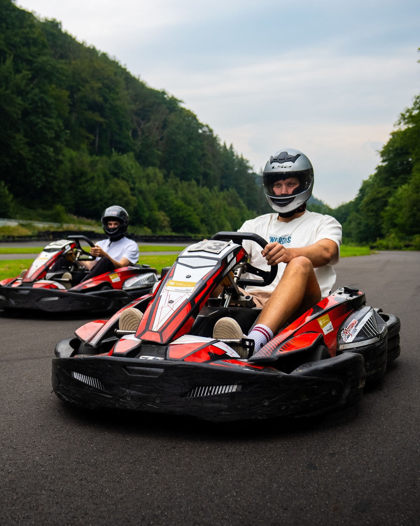Zwei Kartfahrer mit Helmen auf der Outdoor-Rennstrecke des Motodrom Hagen, umgeben von grünem Wald und Asphaltbahn im Naturpanorama