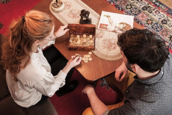 Zwei Personen sitzen an einem Tisch mit einer geöffneten Schatztruhe voller Goldmünzen, neben ihnen ein Rätselheft – Szene aus „Muriel_Ingrid“