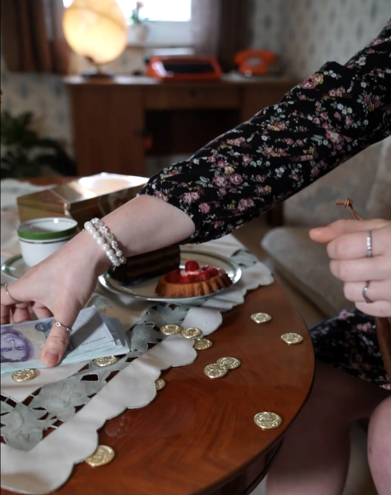 Person greift nach einem Geldschein auf einem Tisch, umgeben von Spielgeld, Gebäck, einer Tasse und nostalgischer Einrichtung – Szene aus „Muriel_Ingrid“