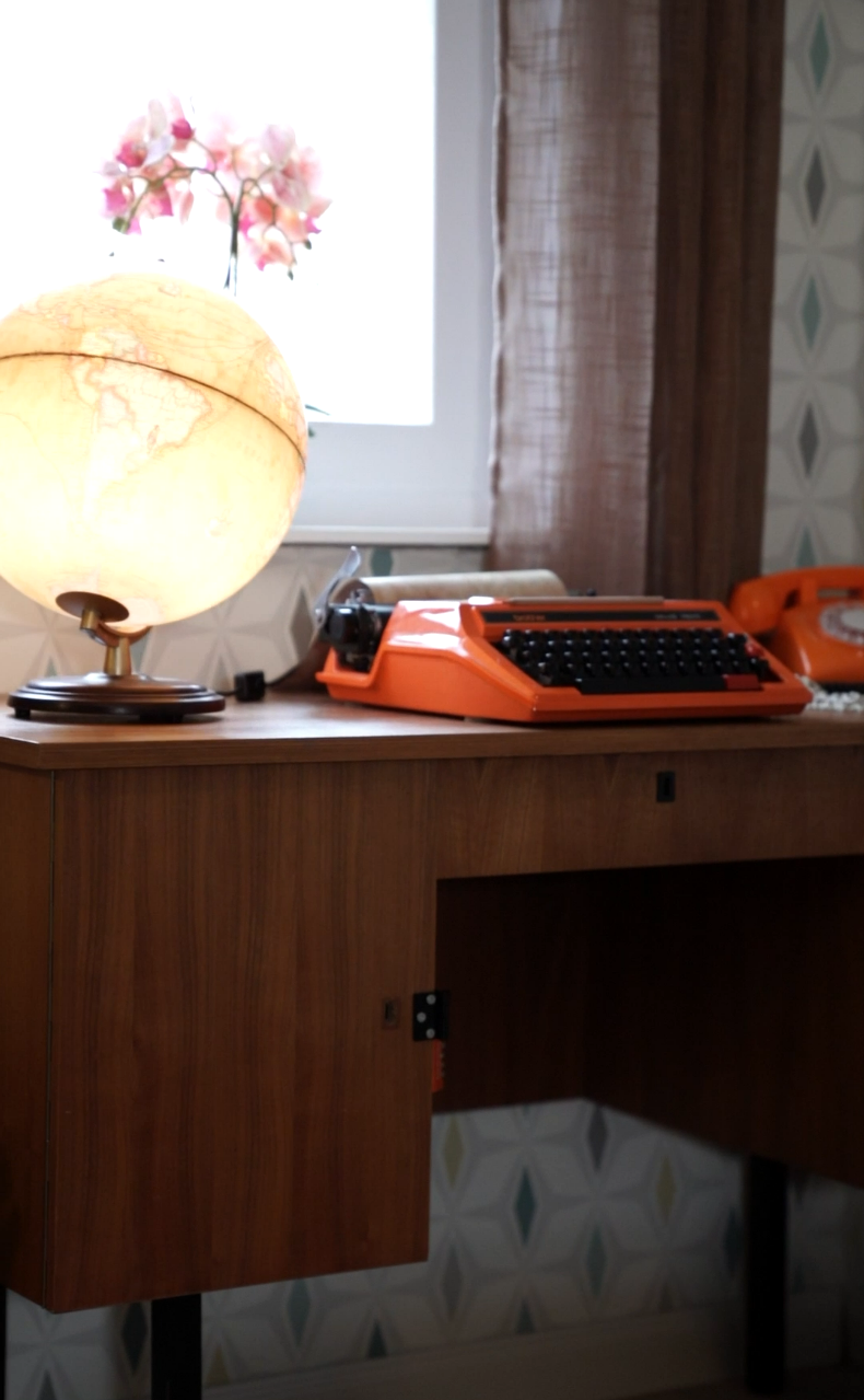 Vintage-Schreibtisch mit oranger Schreibmaschine, passendem Retro-Telefon und beleuchteter Globuslampe vor einem Fenster – Szene aus „Muriel_Ingrid“