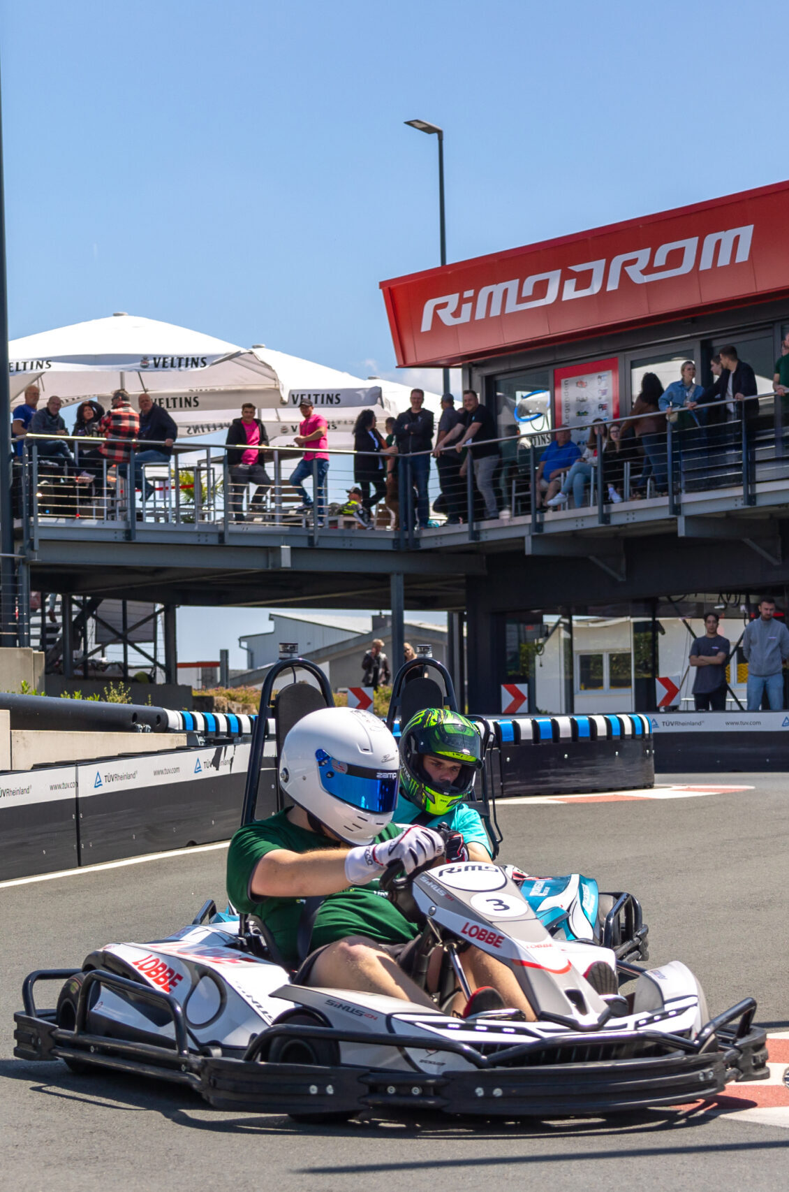 Zwei Fahrer im Doppelkart auf der Outdoor-Rennstrecke des Rimodrom Hemer, im Hintergrund Zuschauer auf der Tribüne unter dem Rimodrom-Schriftzug