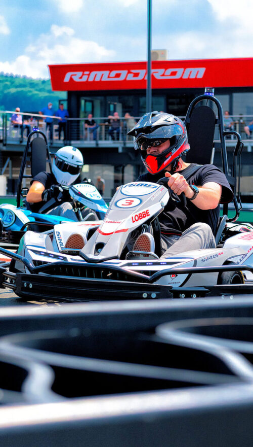 Nahaufnahme eines Fahrers mit Helm im Kart auf der Strecke des Rimodrom Hemer, ein zweiter Fahrer im Hintergrund, Zuschauerbereich und Tribüne leicht unscharf sichtbar