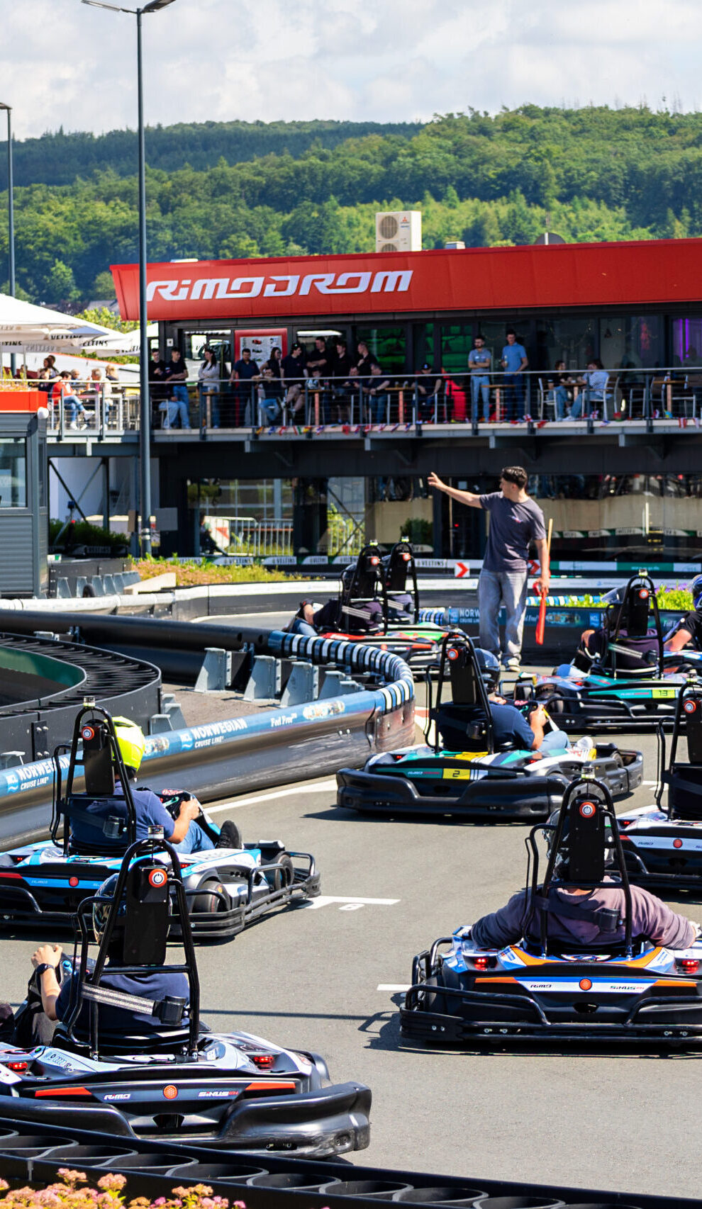 Mehrere Fahrer in Karts an der Startlinie des Rimodrom Hemer, ein Rennleiter gibt das Startsignal, im Hintergrund Zuschauer auf der Terrasse unter dem Rimodrom-Schriftzug