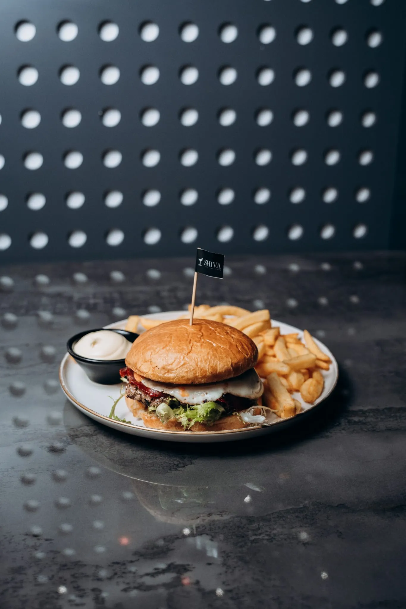 Burger mit Sesam-Bun, Salat, Bacon, Käse und Sauce, serviert mit Pommes frites und Dip auf einem Teller im Shiva Food and More Dinslaken, vor moderner gepunkteter Wand
