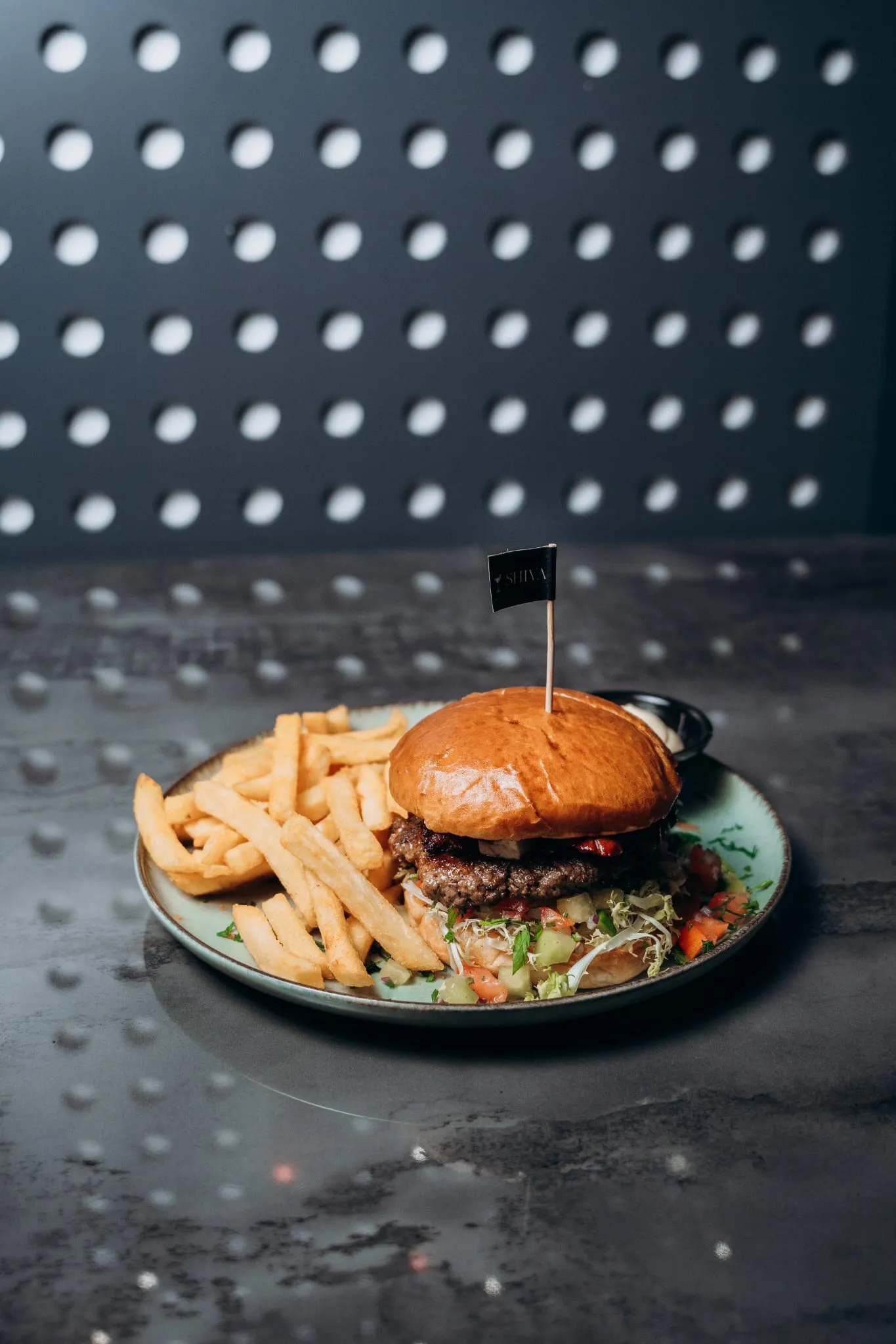 Doppelt belegter Beef-Burger mit Salat im Brioche-Bun, serviert mit Pommes frites und Dip auf einem Teller vor gepunkteter Wand im Shiva Food and More Dinslaken