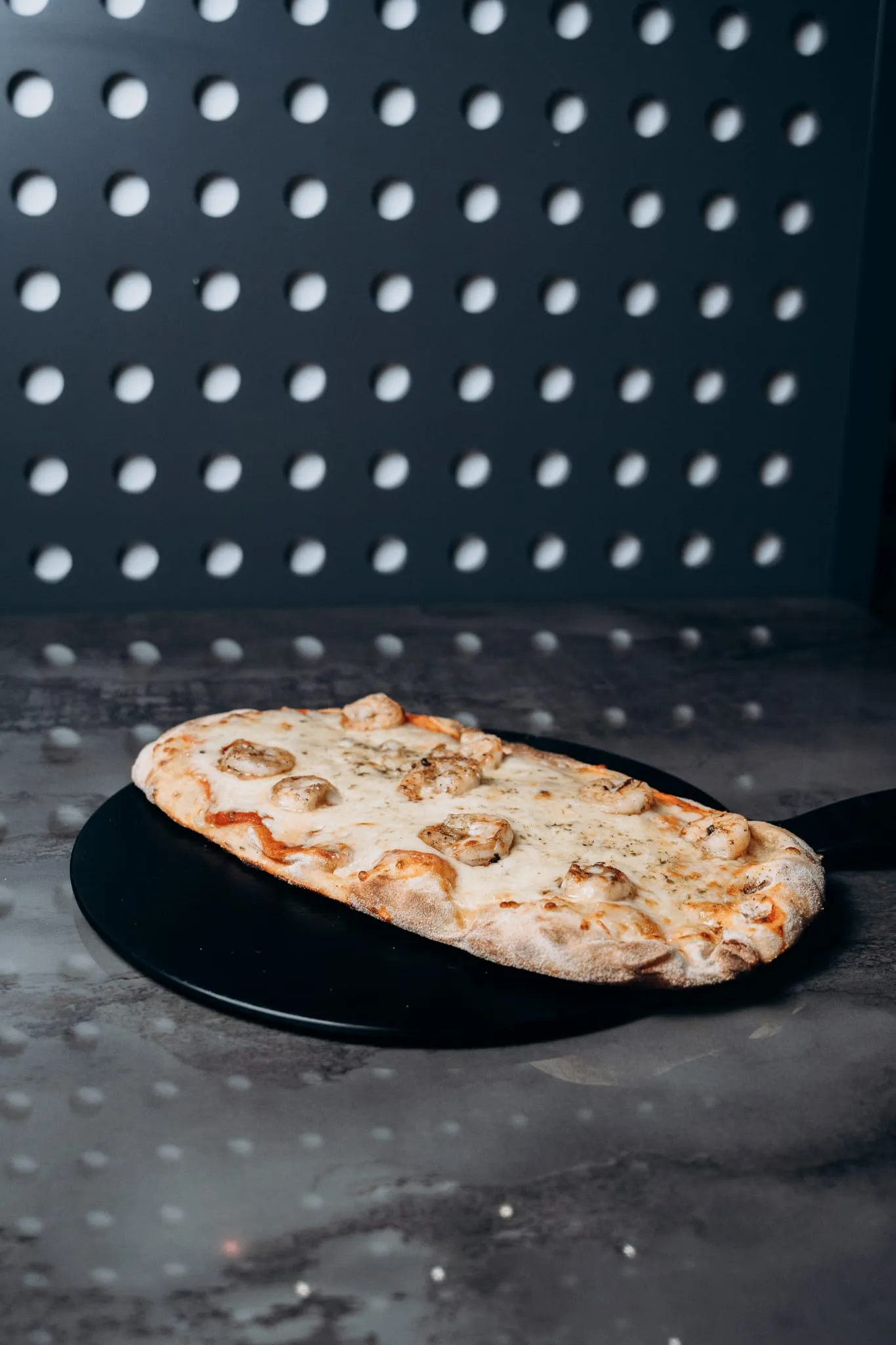 Pinsa mit geschmolzenem Käse und Garnelen, serviert auf schwarzem Teller vor moderner gepunkteter Wand im Shiva Food and More Dinslaken