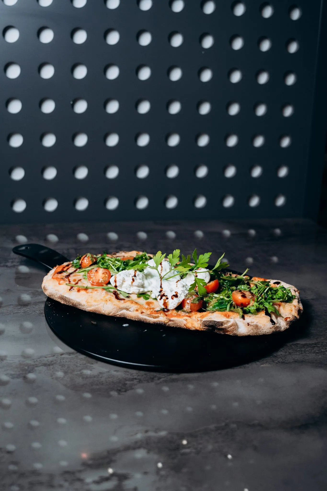 Ofenfrische Pinsa auf dunklem Teller, belegt mit Rucola, Cherrytomaten und einem Klecks Frischkäsecreme – serviert im modernen Ambiente von Shiva Food and More Dinslaken