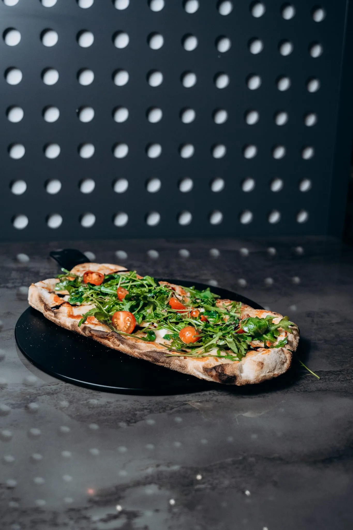 Ofenfrische Pinsa vom Shiva Food and More Dinslaken, belegt mit Rucola, Cherrytomaten und cremiger Basis, serviert auf dunklem Teller vor moderner Wandkulisse