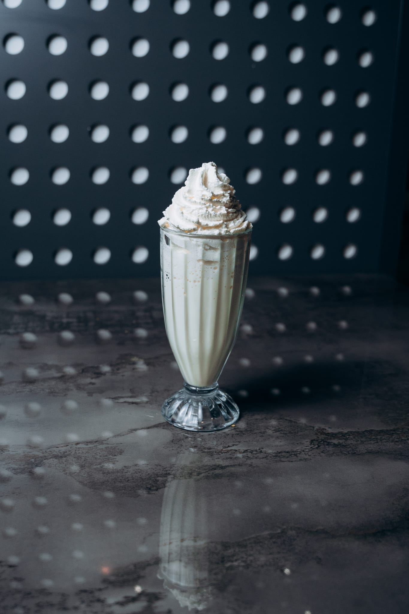 Klassischer Vanille-Milchshake mit Sahnehaube in einem hohen Glas, serviert auf dunklem Tisch vor gepunkteter Wand im Shiva Food and More Dinslaken