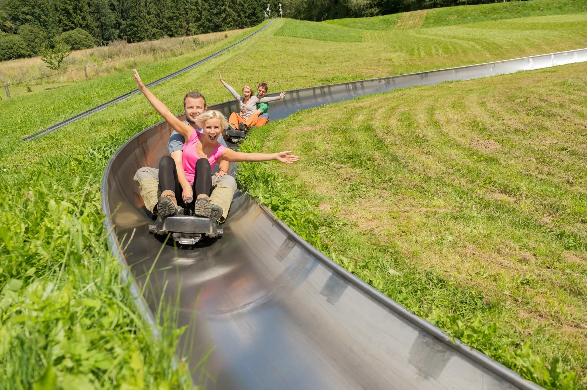 Menschen fahren lachend auf einer Sommerrodelbahn durch eine grüne Wiesenlandschaft