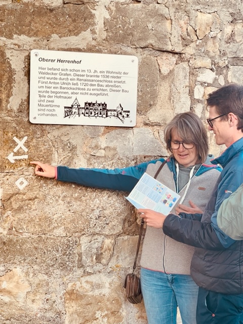 Zwei Stadtkrimi-Teilnehmende analysieren Zeichen an einer historischen Mauer in Kassel, mit Roadbook in der Hand und Infotafel über die Stadtgeschichte im Hintergrund