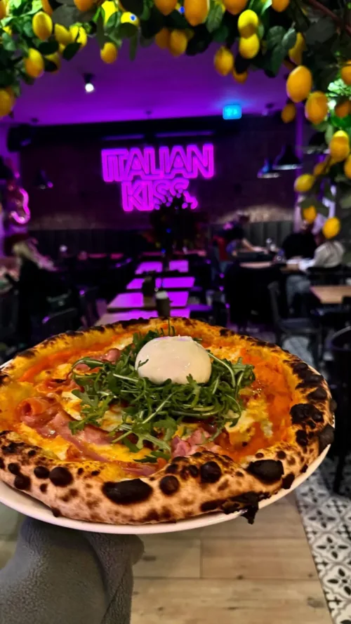 Holzofenpizza mit Burrata, Rucola, Schinken und knusprigem Rand, im Vordergrund serviert, im Hintergrund das pink leuchtende „Italian Kiss“-Neonlogo im Restaurant