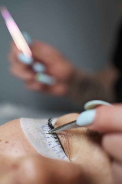 Nahaufnahme einer Wimpernverlängerung mit Pinzette, bei der einzelne Extensions auf die Naturwimpern appliziert werden, das Auge ist mit einem Schutzpad abgeklebt