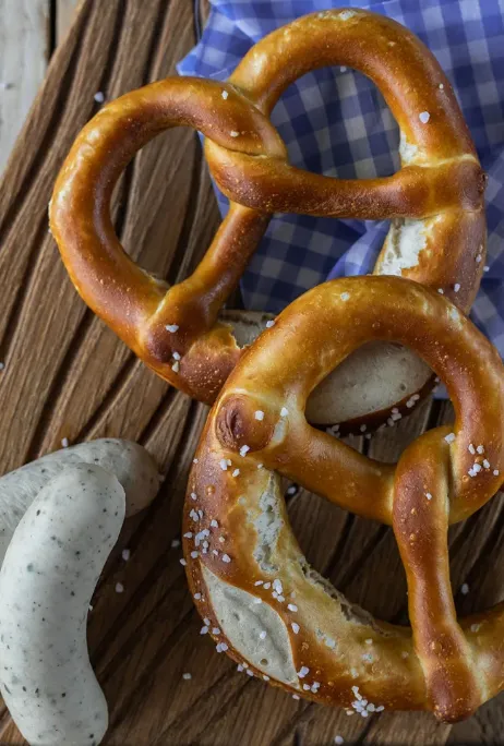 Frische Brezeln und Weißwürste auf einem Holzbrett mit blau-weißem Tuch – typische Brotzeit in der Alm Fernweh