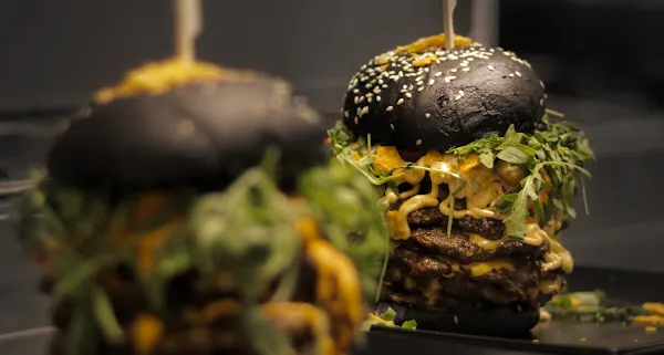 Doppeldecker-Burger mit schwarzem Sesam-Bun, mehreren Fleischpatties, Käse, Salat und Zwiebeln