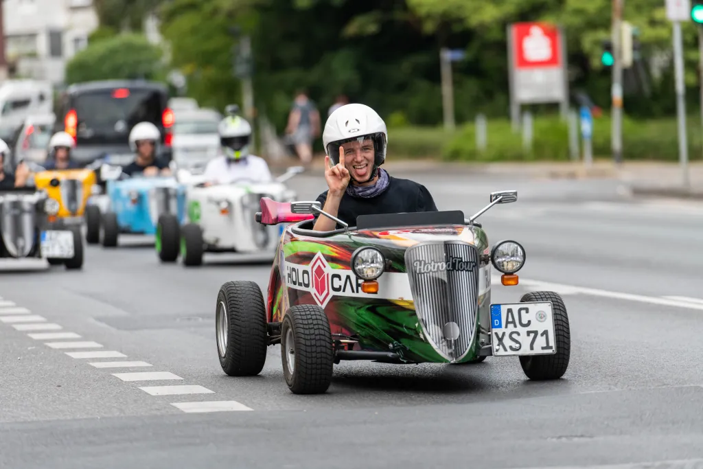 Teilnehmer der Hotrod Tour Aachen fährt lachend mit einem Mini-Hotrod durch die Innenstadt, gefolgt von weiteren Fahrern im Konvoi