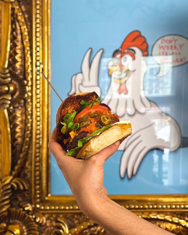 Veganes Burger-Highlight von Lia’s Kitchen in Berlin – in der Hand gehalten vor humorvollem Hühner-Motiv